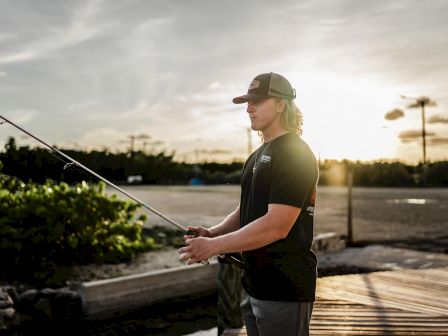 A person wearing a cap is fishing at sunset near a dock with greenery visible in the background and a partly cloudy sky.