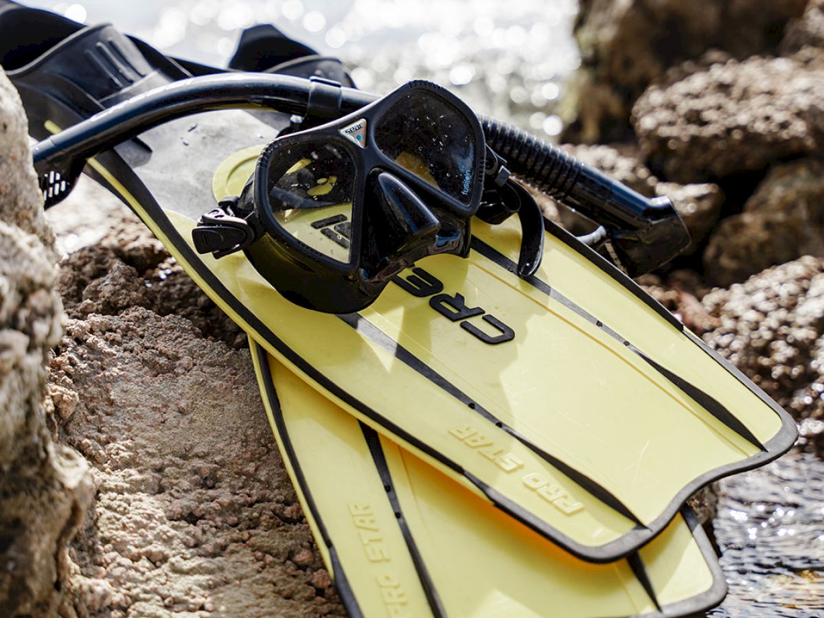 The image shows yellow snorkel fins, a black snorkel, and a diving mask resting on a rocky surface by the water.