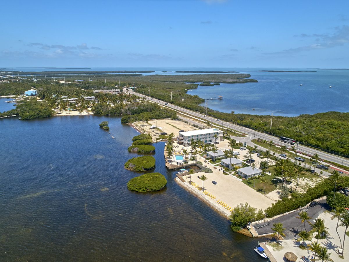 An aerial view of a coastal area featuring a roadway, buildings, docks, and surrounding water bodies with lush greenery and small islands.