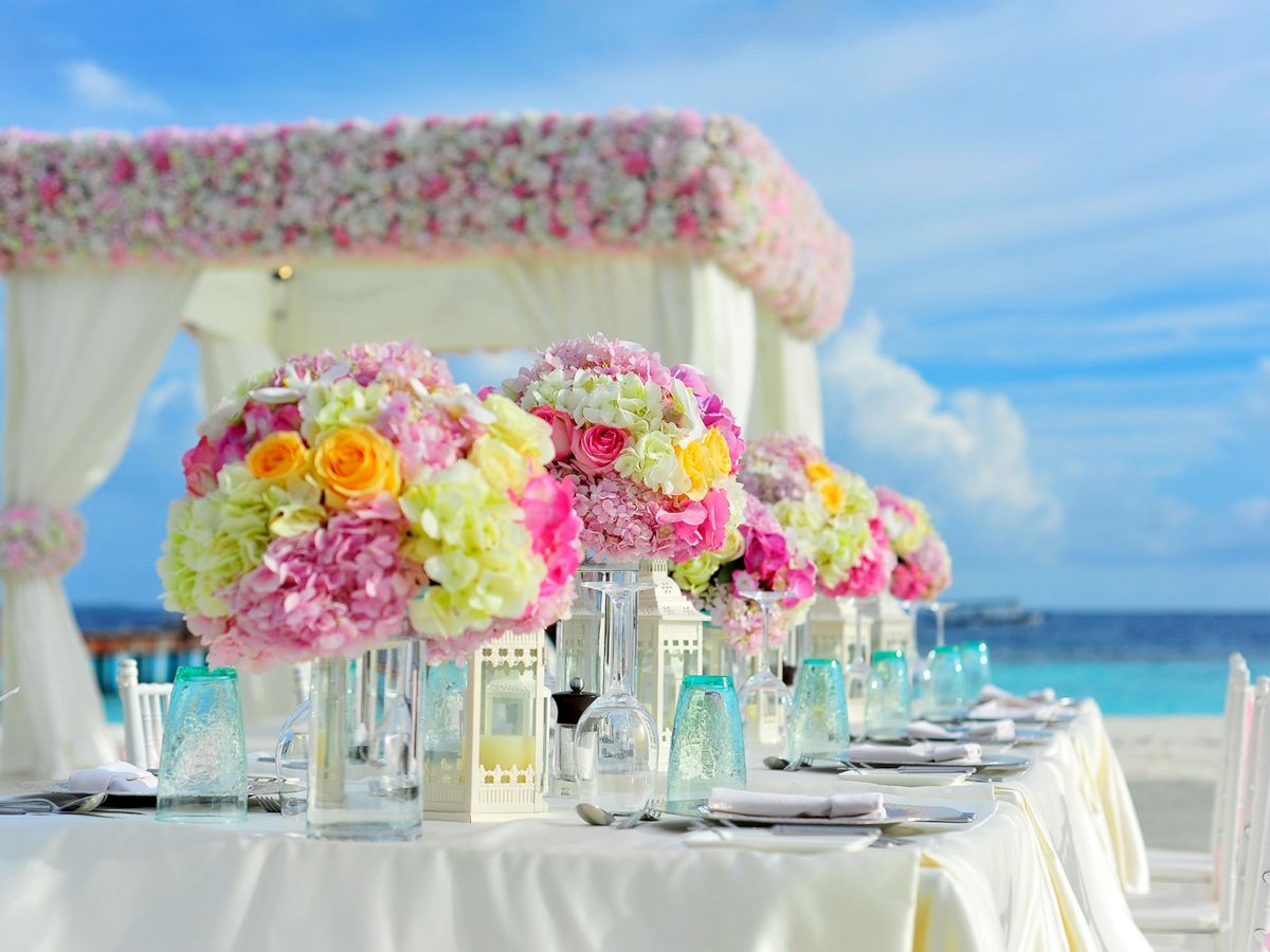 A beautifully set table with colorful floral centerpieces under a flower-adorned canopy by the seaside, ideal for a picturesque event or wedding.