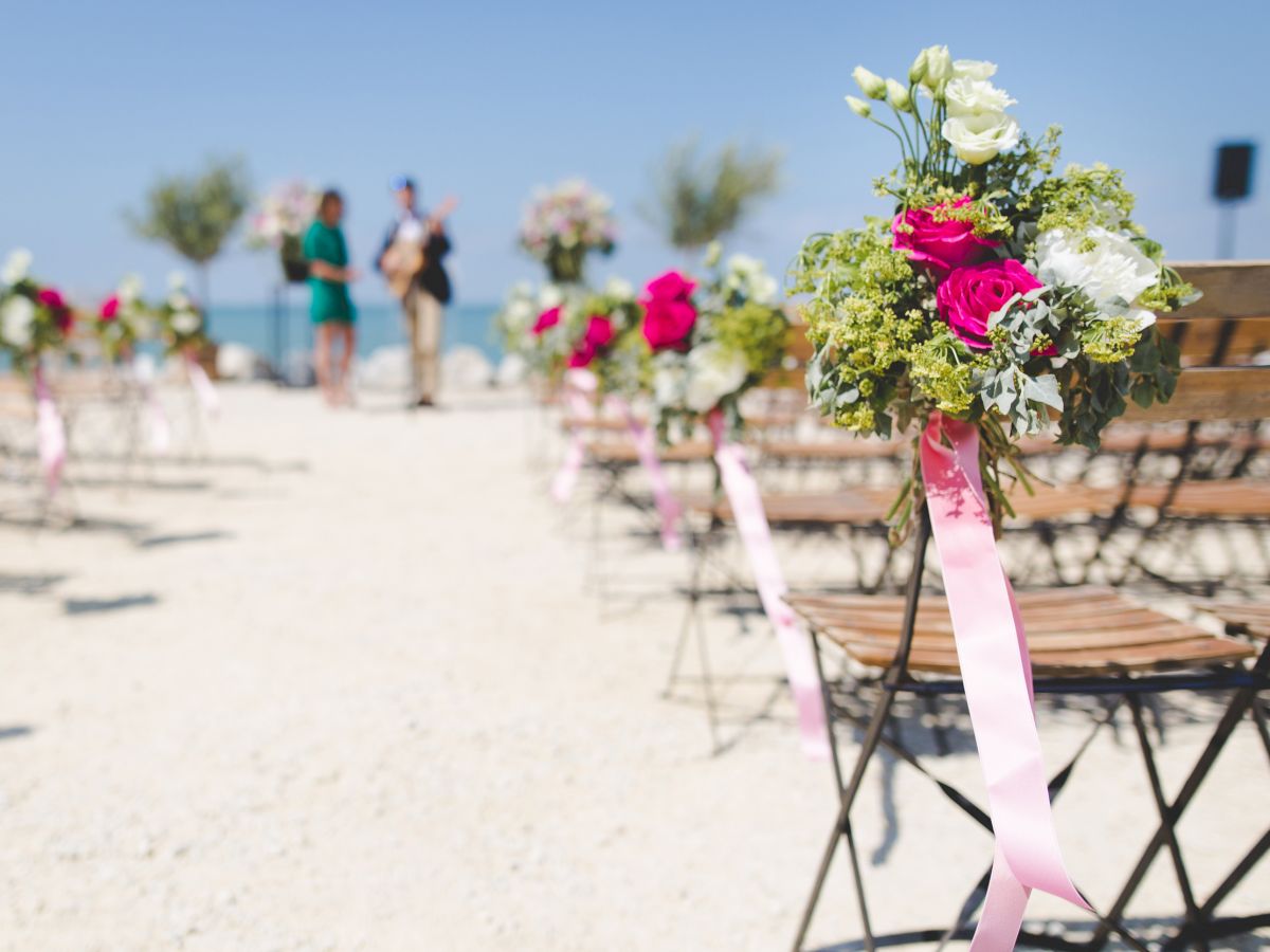 Outdoor wedding setup with chairs adorned with floral decorations and pink ribbons, leading towards an altar with two blurred figures in the background.