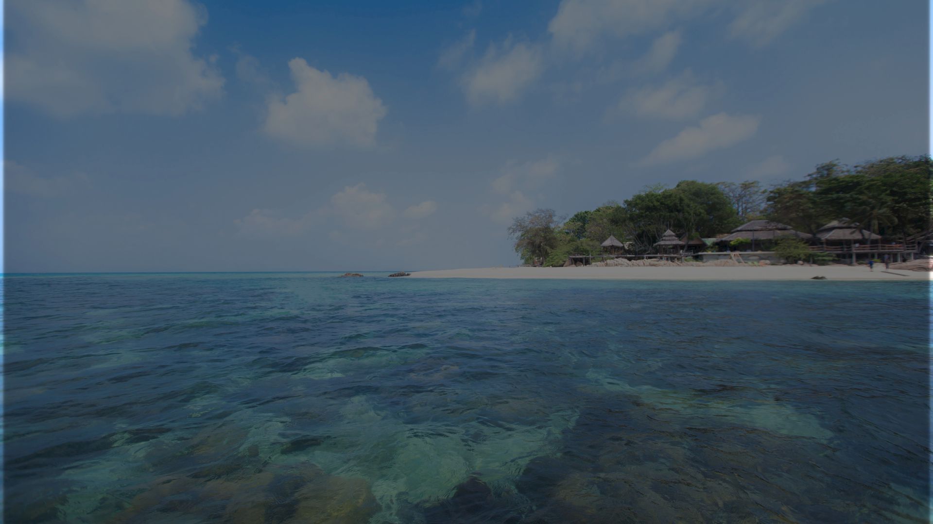 A serene beach scene with clear, turquoise water, a distant shoreline with some trees and huts, and a partly cloudy sky.