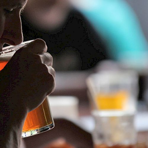 A person is drinking from a glass of beer in a dimly lit setting with other people and drinks in the background.
