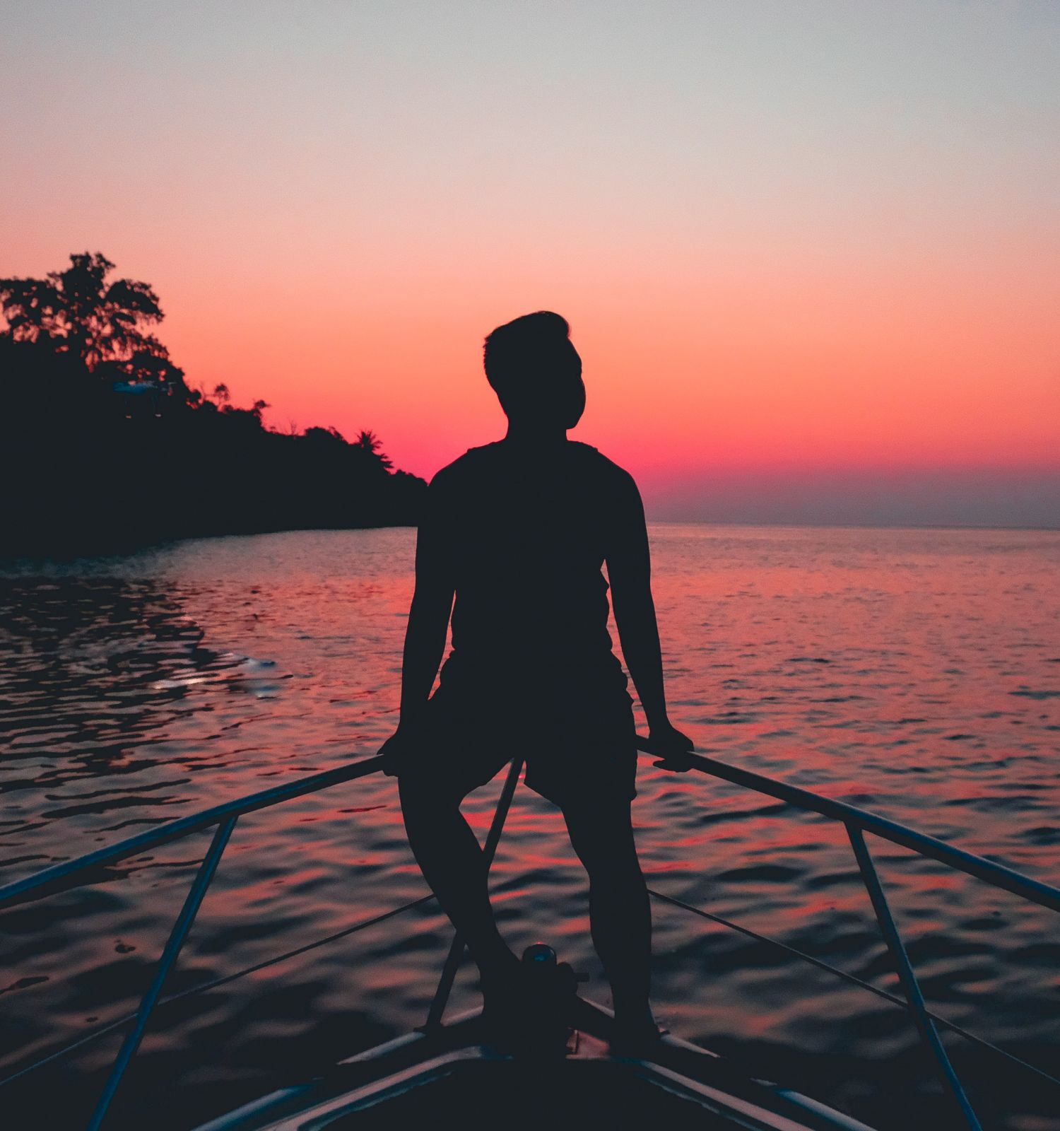 A person is standing on the bow of a boat, silhouetted against a vibrant sunset over the water with a distant shoreline visible.