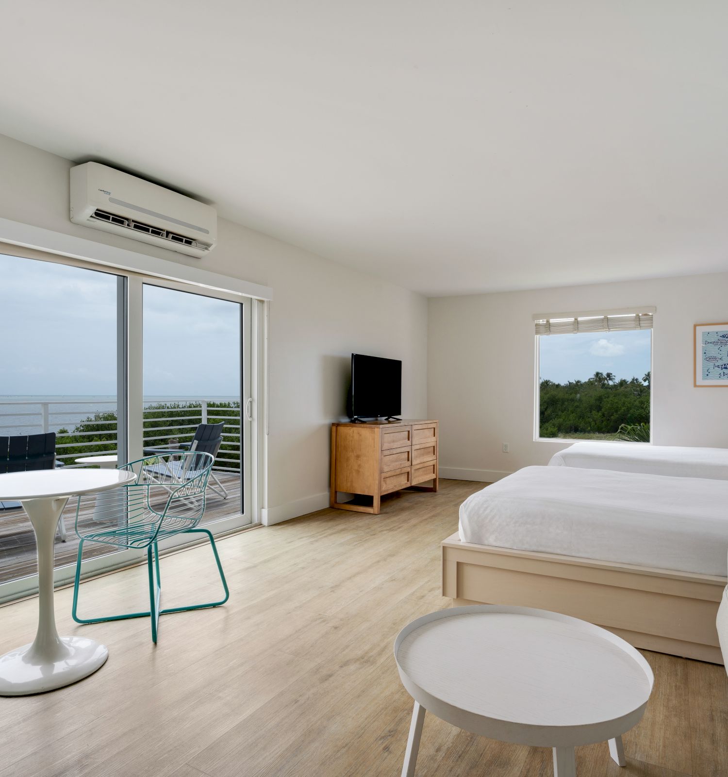 A modern hotel room with two beds, a couch, a TV, and a balcony overlooking the sea; features minimalist furniture and a blue accent wall.