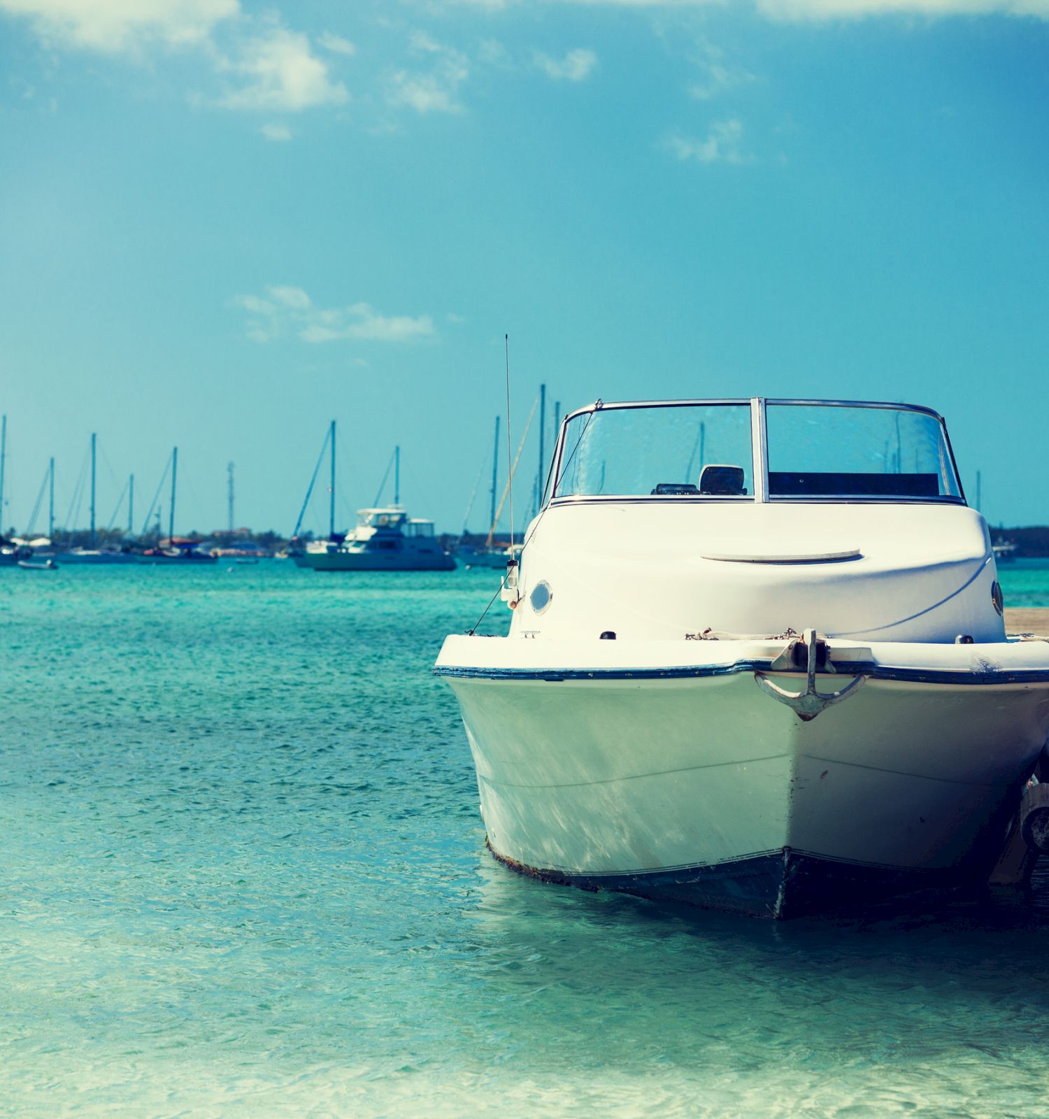 A white motorboat is docked at a pier over clear turquoise water with a background of numerous sailboats.