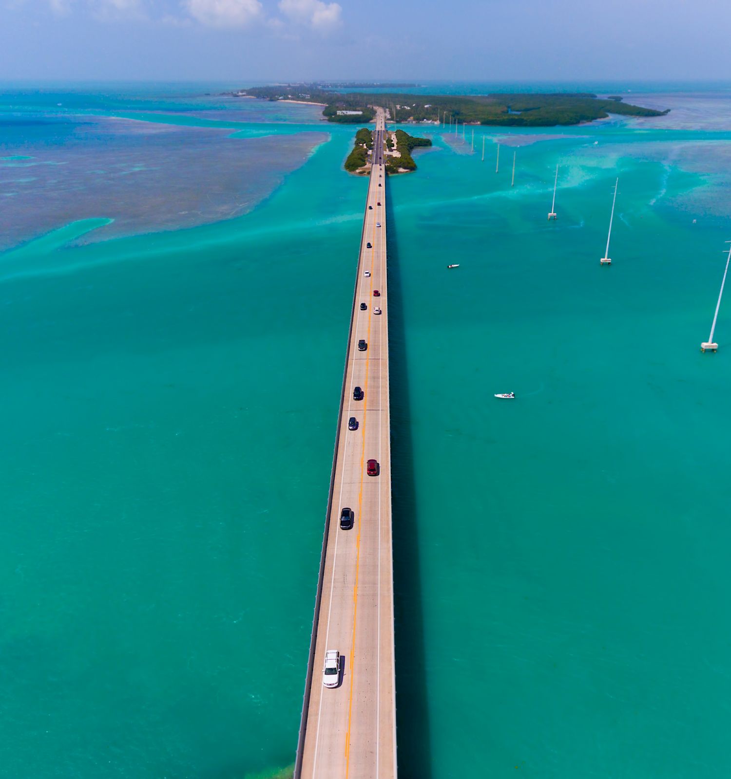 A long, straight bridge stretches across vivid turquoise water, with several vehicles traveling on it, leading towards an island in the distance.